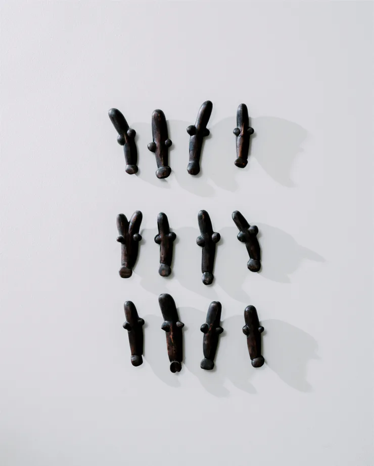 Sixteen black cylindrical metal objects arranged in four rows against a white wall, casting distinct shadows, installation by Minjae Kim.