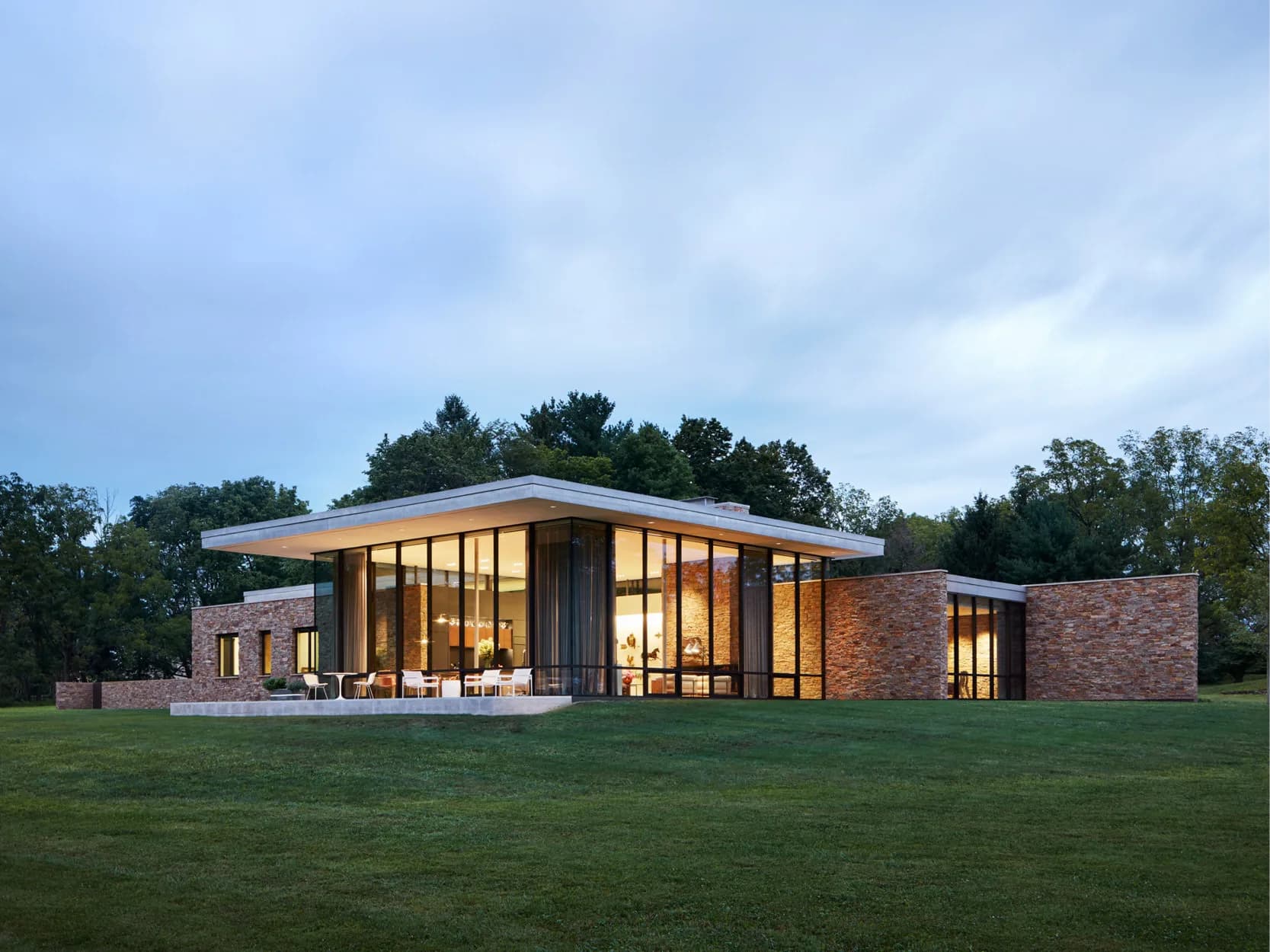 Modern brick and glass residential building with illuminated interior, expansive lawn, and natural landscape backdrop by Stephen Cassell.