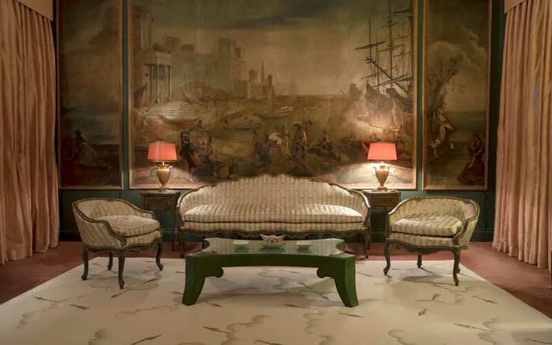 Elegant living room with striped settee and matching chairs flanking a green bench, accented by table lamps and large classical mural backdrop by Charlap Hyman & Herrero.