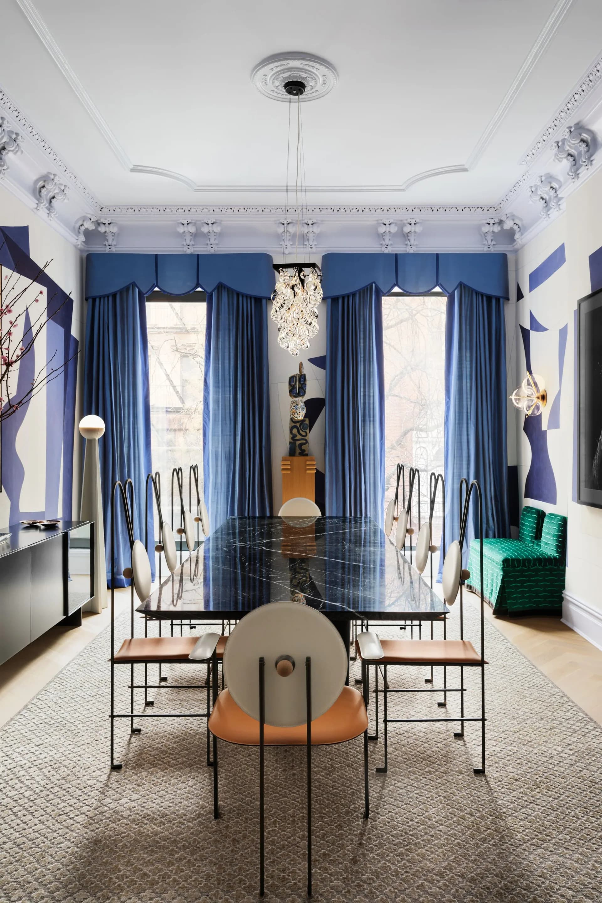 Elegant dining room featuring blue velvet curtains, black glass table with modern chairs, crystal chandelier, white coffered ceiling, and blue accent walls by Danielle Colding Design.