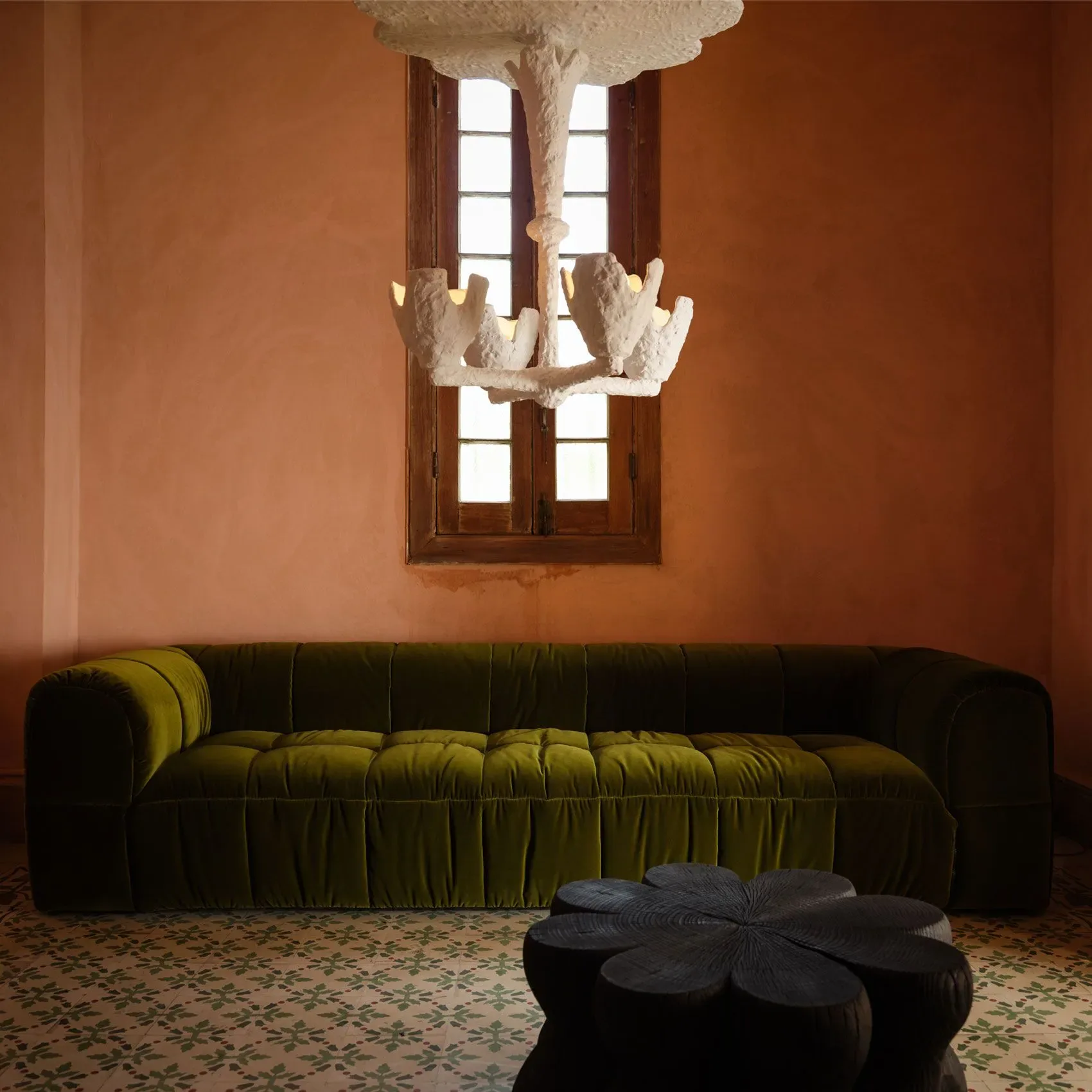 Terracotta-walled room with pleated olive-green sofa, sculptural white plaster chandelier, and black organic ottoman on patterned floor by The Future Perfect.
